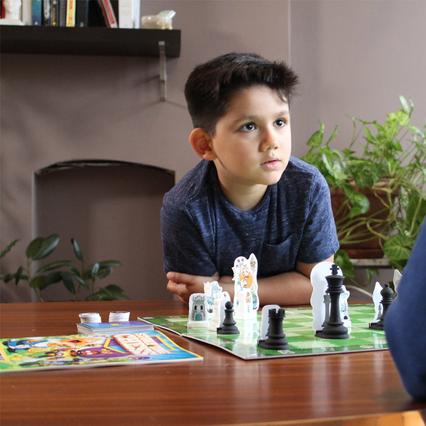 A dark-haired boy sits at a table with the Story Time Chess board in front of him. He is looking up at someone across the table, but you can only see a part of this persons left arm. The game board on the table has light and dark checkered squares with the game pieces in play. To the left you can see the story book and instructions and some playing cards with round token pieces.