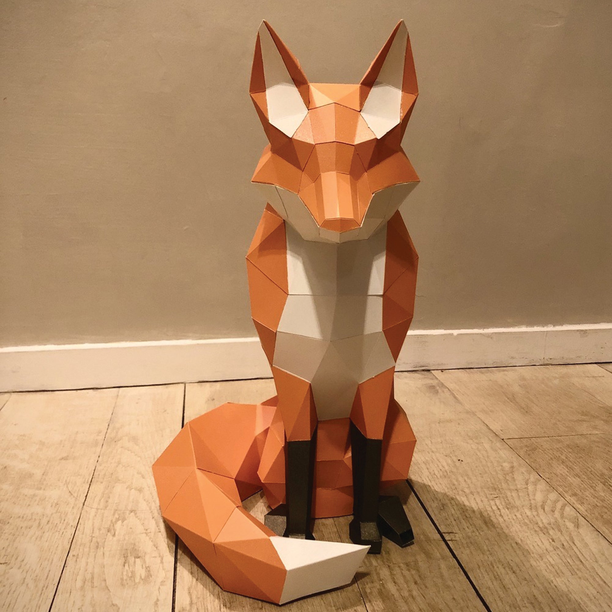 Papercraft fox with a dark beige wall behind and a wood floor underneath. Geometrical-shaped fox that is folded and cut paper pieces glued together. Fox is mainly a dark orange with black paws and a white chest, chin area, inside ears, and tip of tail.