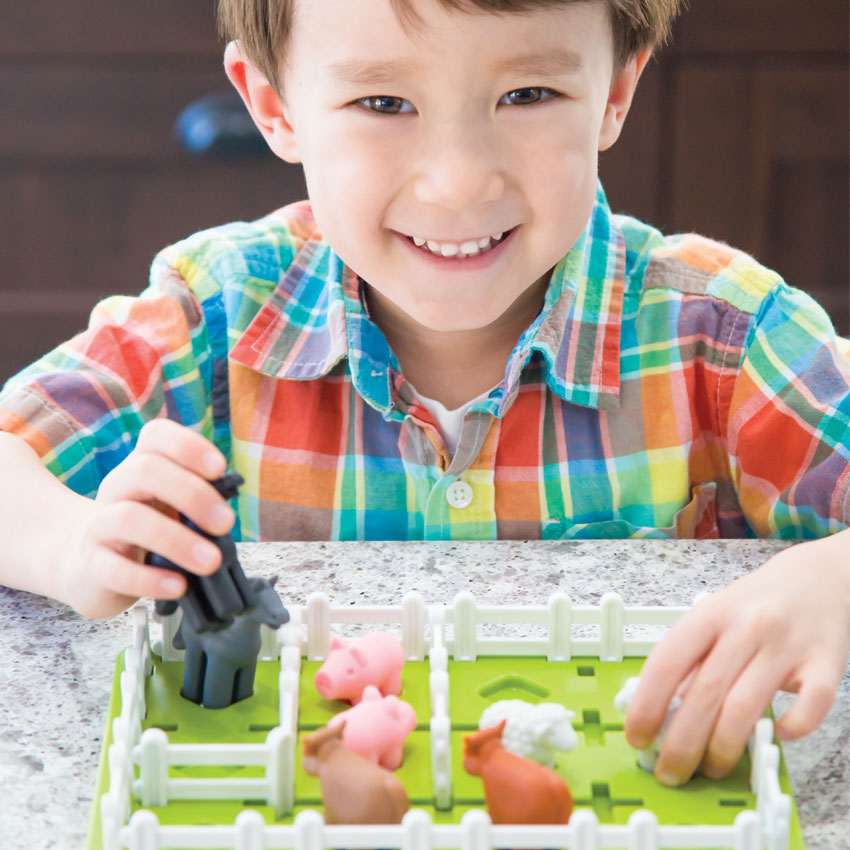 A young brunette boy, smiling, and playing with the Smart Farmer game. He is holding a horse piece with his right hand and grabbing onto a sheep piece with his left. The game board is green and rectangular with white fence pieces all around the outside. Inside are a few more fence pieces, a horse, pigs, cows, and sheep.