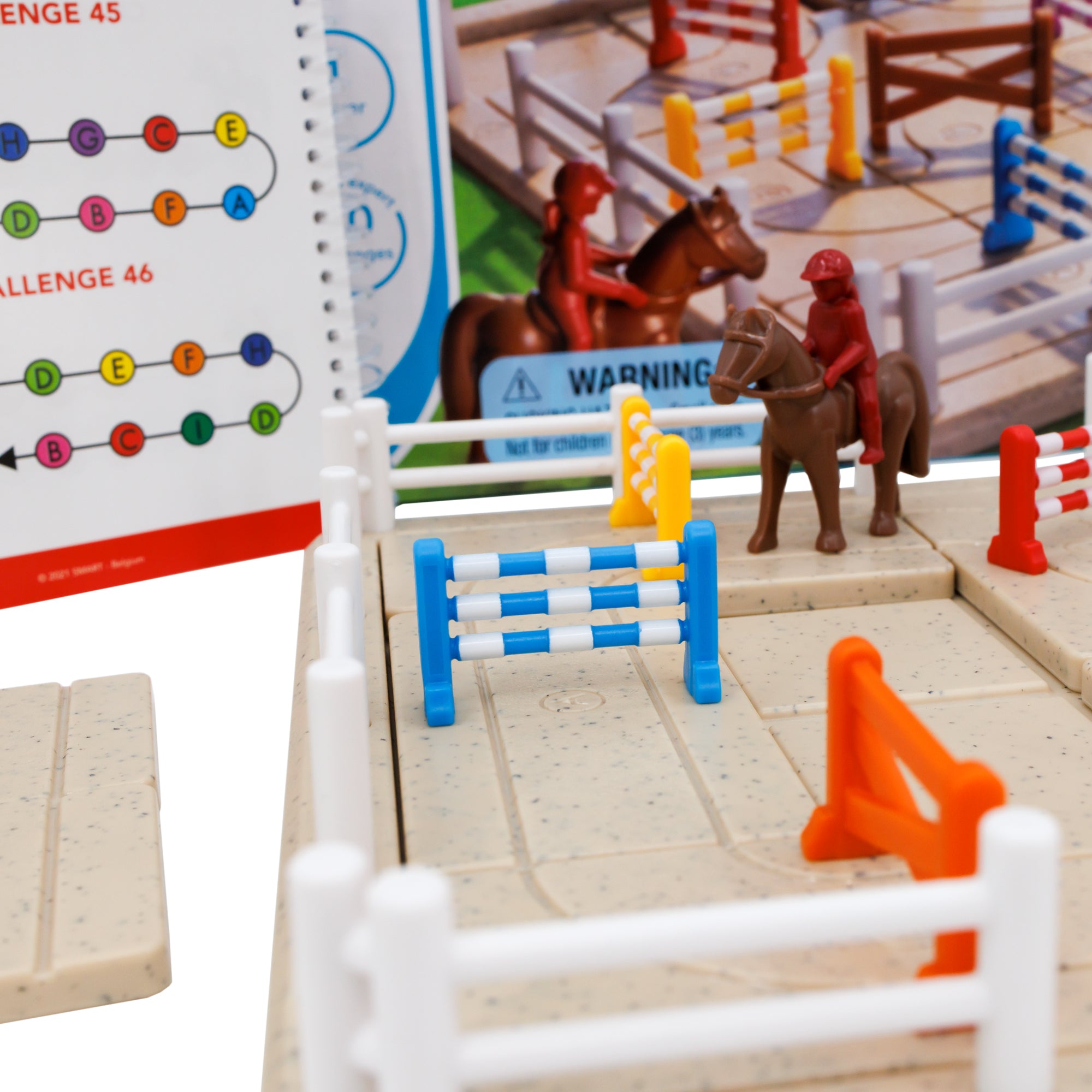 A close up of the Horse Academy game. The game board is a sand-colored rectangle with white fence pieces around the edges. There a several jumping fence pieces in many colors on the game board and a red girl character on top of a horse piece in between the fences. You can see the game box and the instruction booklet in the background, out of focus.