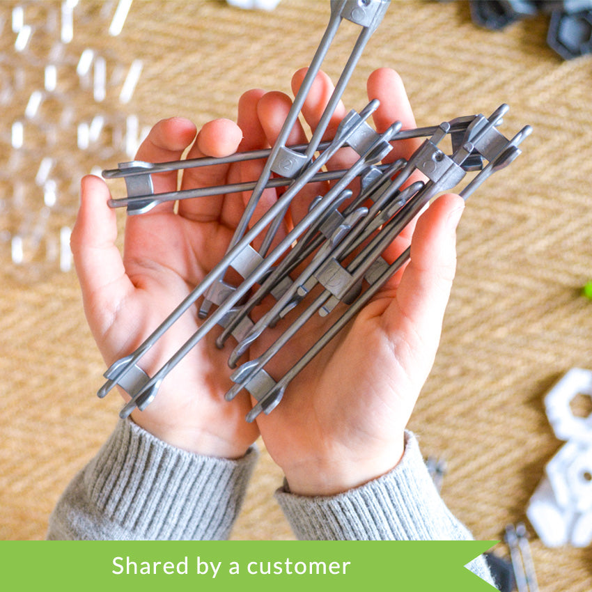 A customer photo from the top of a child’s hands holding several gray rail pieces from the GraviTrax set. Below the hands, and out of focus, are playing pieces from the set. The pieces shown are white hexagons, green hexagon attachment pieces, gray hexagons, and clear hexagon plates.