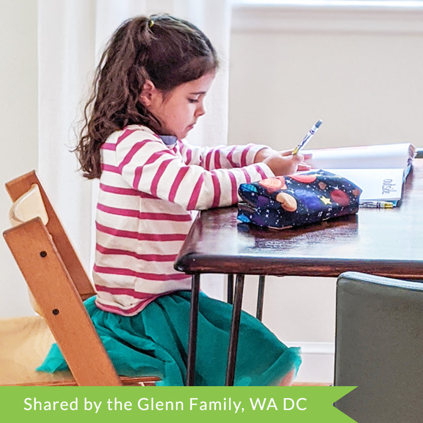 A customer photo of a brunette girl in a striped shirt and green skirt sitting at a table, drawing with a coloring tool in the I Can Doodle Rhymes book.  A space themed pencil case sits on the table blocking out a lot of the book.