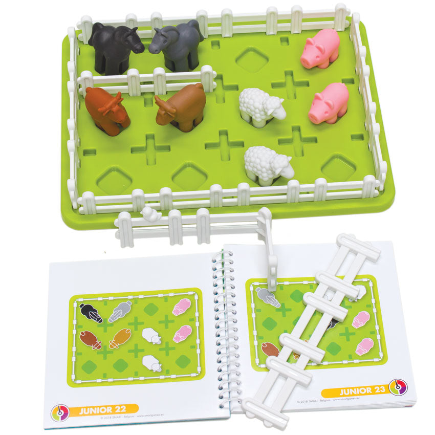 Smart Farmer game components. The game board is green and rectangle-shaped with white fence pieces all around the edge. You can see pigs, sheep, cows, and horses inside the fence. Off to the side are more fence pieces laying on top of the instruction booklet, open to show 2 junior challenges.