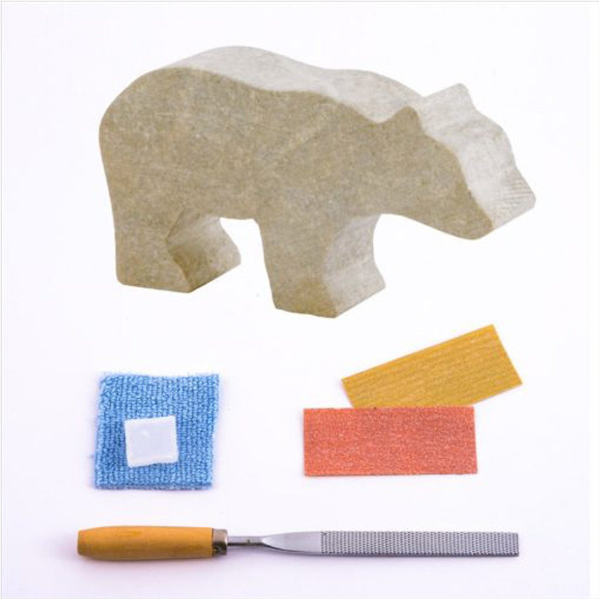 Bear Soapstone Carving Kit contents. On top is a block-like bear shaped soapstone. Under that  are red and yellow sanding pieces and a  blue cloth with a square chunk of wax on top. On the bottom is a metal file with a wood handle.