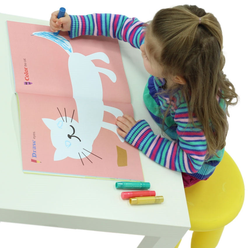 Draw + Learn Animals + Places book is open to show a white cat shape on a pink page. A girl, sitting on a yellow plastic stool, is wearing a striped shirt and is coloring the cat's tail blue. To the left of the girl, on the white table, are 3 coloring tools in yellow, pink, and green.