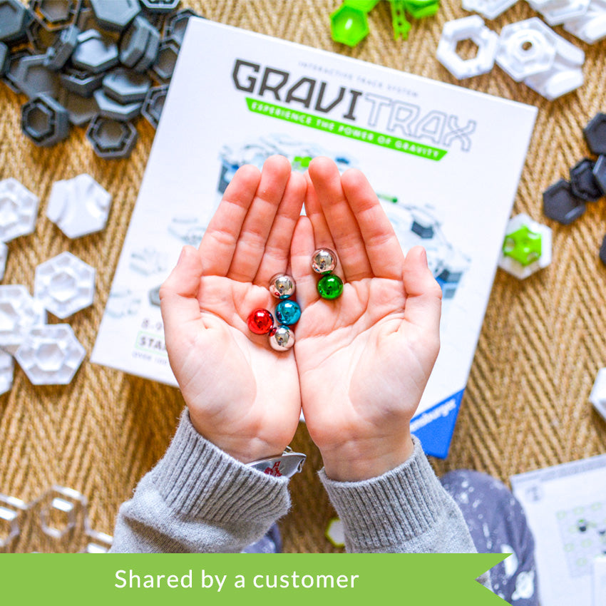 A customer photo from the top of a child’s hands holding the red, green, blue, and silver marbles from the GraviTrax set. The box is below the hands, out of focus, and pieces are scattered all around the box. The pieces are dark gray hexagons, white hexagons, green hexagon attachment pieces, and gray rails. 