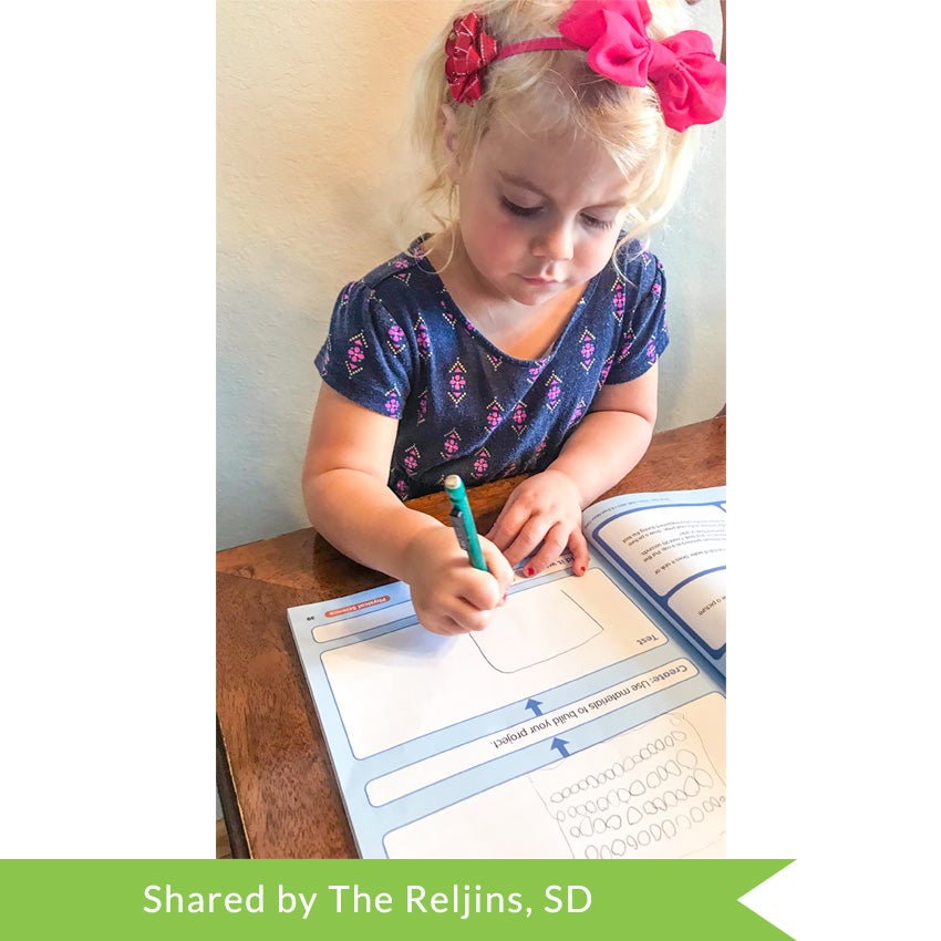 A customer photo of a young blonde girl with a large pink bow in her hair, drawing a picture in the Smart Start Stem Pre K book. The open book pages are white and blue. She is in the middle of drawing a large square.