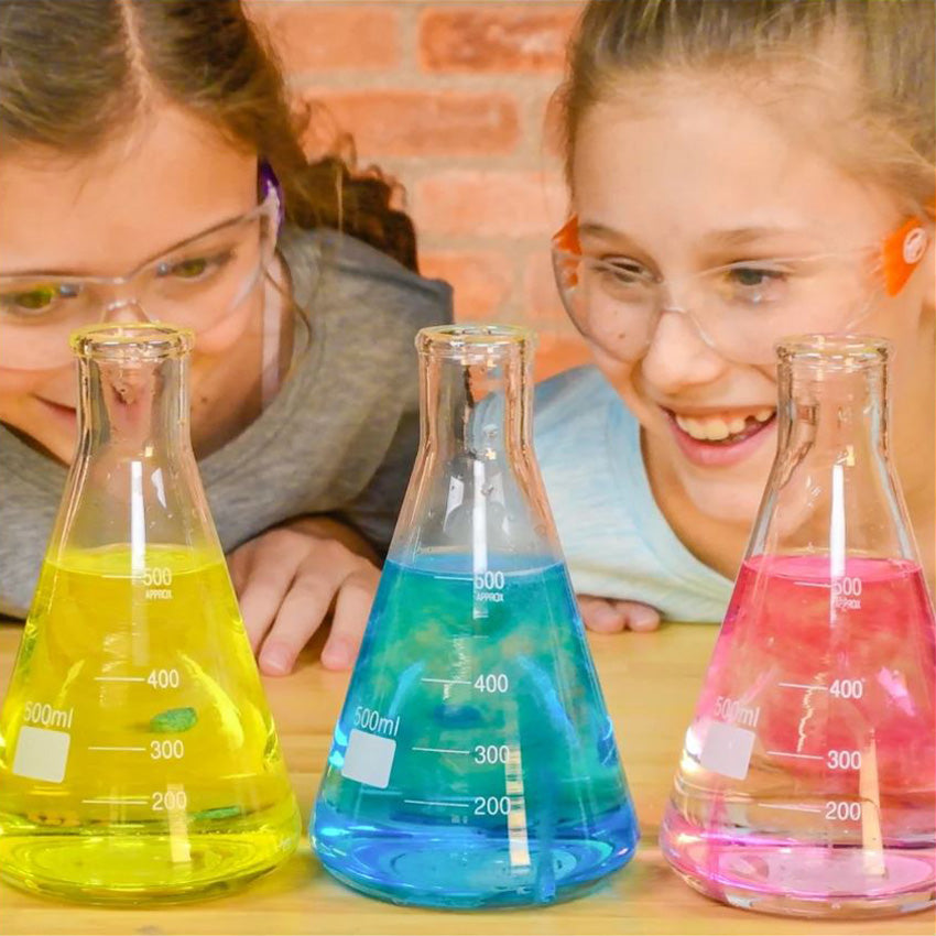 Two girls with safety goggles watch with smiles as Color Fizzers color beakers with water in front of them. The left beaker has yellow water with a yellow tablet coloring the water. The middle beaker has blue water with a blue tablet coloring the water. The right beaker has pink water with a red tablet coloring the water.