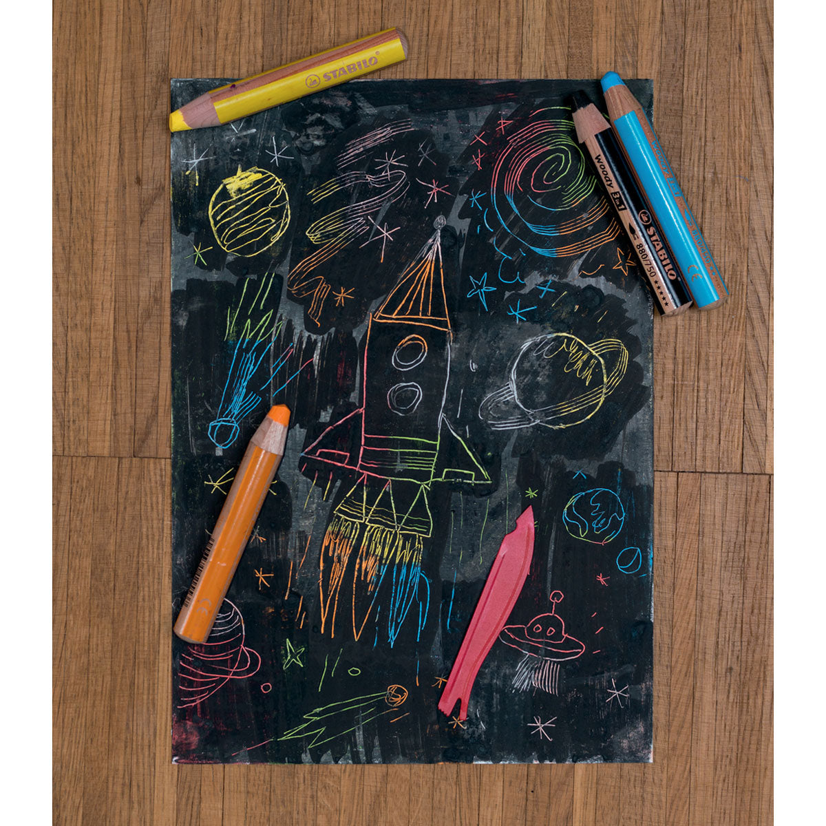 Wooden floor background with a black chalk picture of  a rocket in space with stars, planets, comets, and a UFO. A variety of colors come through the scratched canvas paper. Four Stabilo pencils in yellow, orange, blue, and black placed on top of the picture along with a scraping tool.