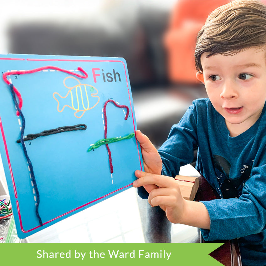 A customer photo of a brunette boy in the middle of talking and holding up a Wikki Stix Alphabet card. The card is blue with an upper and lower case F along with a fish on it. The wax sticks he used to cover the letters are blue, pink, green, and purple.