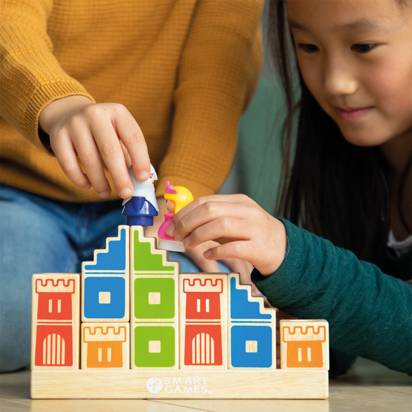 A young dark-haired girl and a boy are playing with the Camelot Junior game on the floor. The boy is is holding a knight piece on top of wood blocks and the girl is holding the princess piece over wood blocks. The base is a long, rectangle wood piece. The wood block pieces, stacked on top, have castle walls painted on them in different colors.
