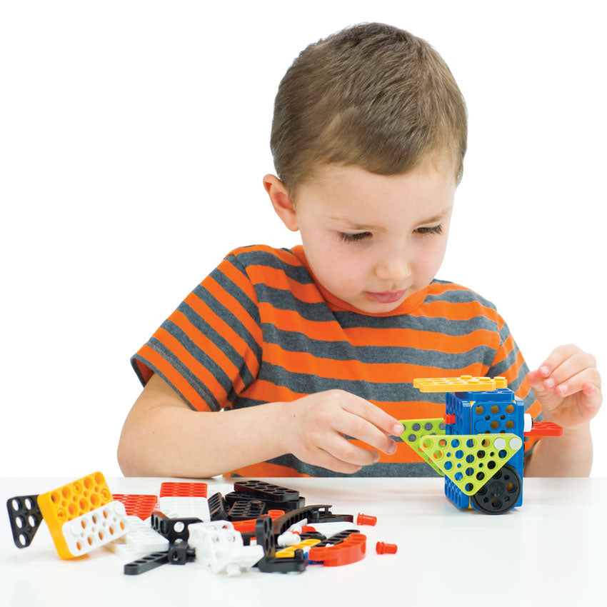 A young boy in a striped shirt is putting together a bluebird robot with other Robotis Play 600 Pets pieces scattered on the table in the bottom-left of the picture.