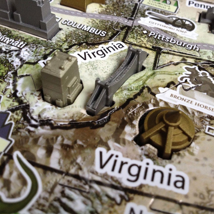 A close up of a the U S A History Over Time Puzzle. The section focuses on Virginia and West Virginia. There are gold, silver, and bronze colored buildings. The puzzle base shows major highways and different types of ground. You can see Pittsburgh with an image of a cannon in the top part of the picture.