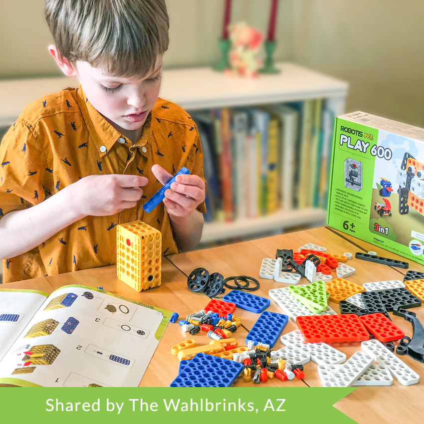 A customer photo of a blonde boy in an orange shirt with dinosaurs putting together a Robotis Play 600 Pets figure. On the table in front of him are many scattered pieces and the instructions book open while he puts a peg in a blue piece.