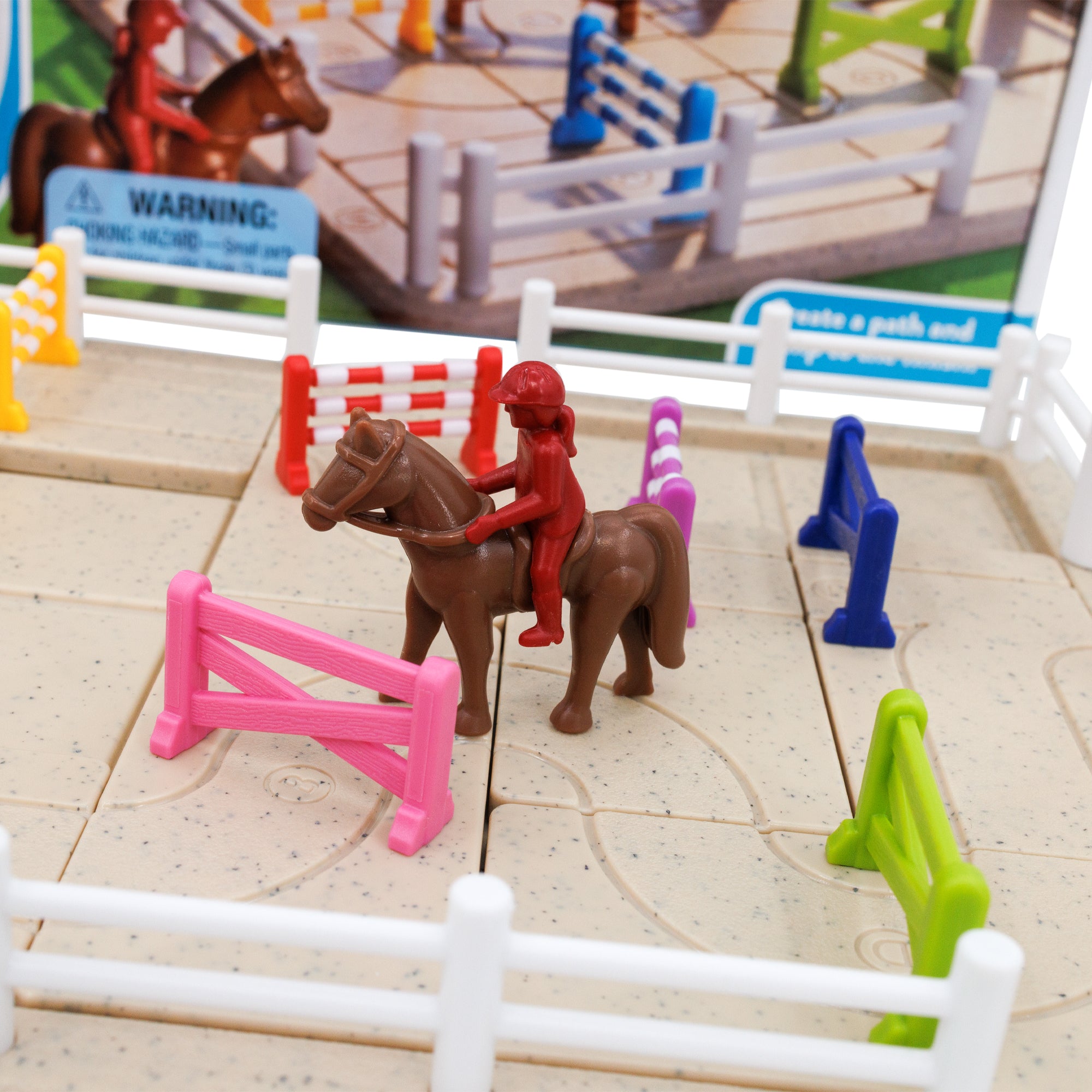 A close up of the Horse Academy game. The game board is a sand-colored rectangle with white fence pieces around the edges. There a several jumping fence pieces in many colors on the game board and a red girl character on top of a horse piece in between the fences. You can see the game box in the background, out of focus.