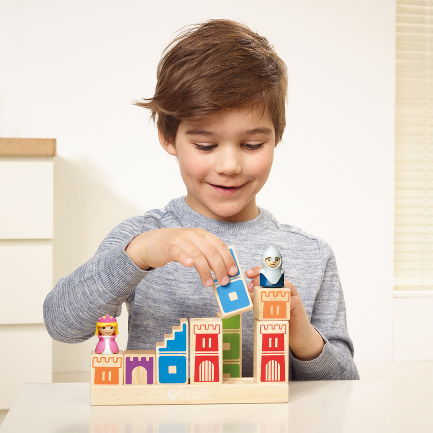 A young brunette boy, smiling, and playing with the Camelot Junior game. He is placing a blue painted wood piece onto the wood base. There is a princess piece standing on a block on the left and a knight piece standing on wood block to the right. The wood block pieces have castle walls painted on them in different colors. The game base is a long, wooden, rectangle with blocks stacked on top.