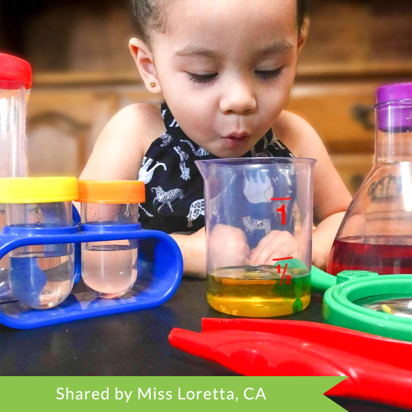Customer photo of a brunette girl wearing a black shirt with white African animals. Her mouth is in the position of saying woah as she is looking into a measuring cup with yellow liquid and a green coloring tablet in the process of coloring the liquid green.  On the table in front of her are other contents of the Primary Science Lab Kit including test tubes in stands, large red tweezers, a green magnifying glass, and a beaker with dark red liquid inside.