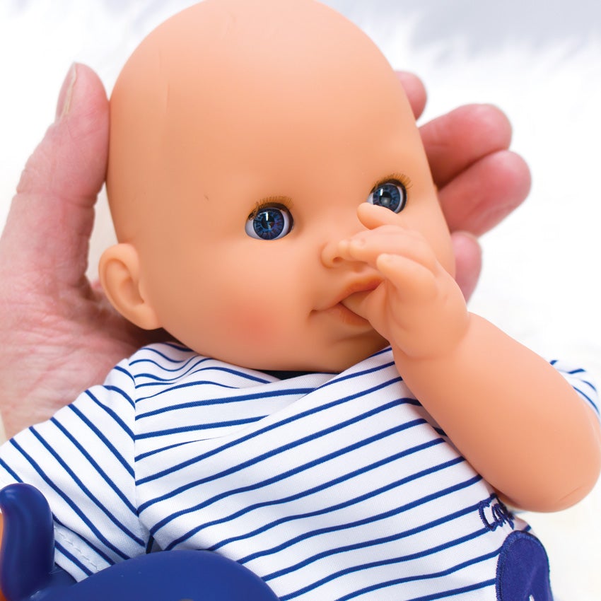 A close up shot of Corolle’s Bebe Marin’s head being held up by a person’s hand. The doll’s arm has been placed, so he is sucking his thumb. He is light skinned with blue, blinkable eyes. He is wearing a white with blue stripes.