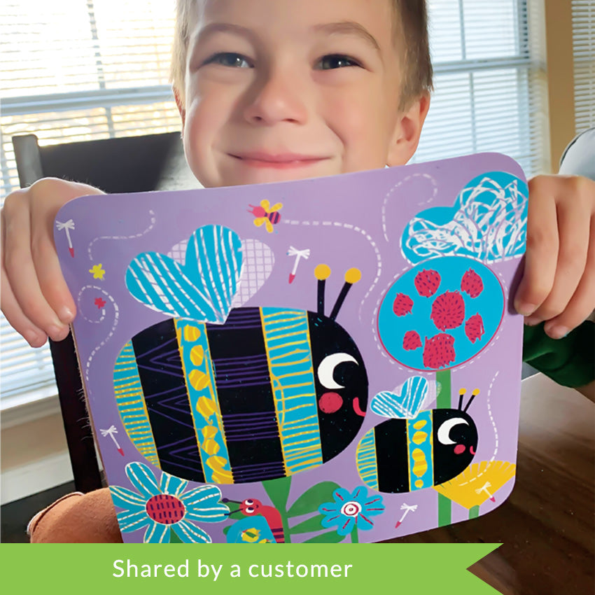 A customer photo of a dark blonde boy smiling big while holding up one of the Djeco Scratch Boards projects. The background is lavender with 2 bees. One large one in the middle-left and one smaller one in the bottom-right. The bees are black, blue, and yellow with patterns scratched onto them. There are flying bugs and flowers throughout the image and a snail climbing up a flower stem.