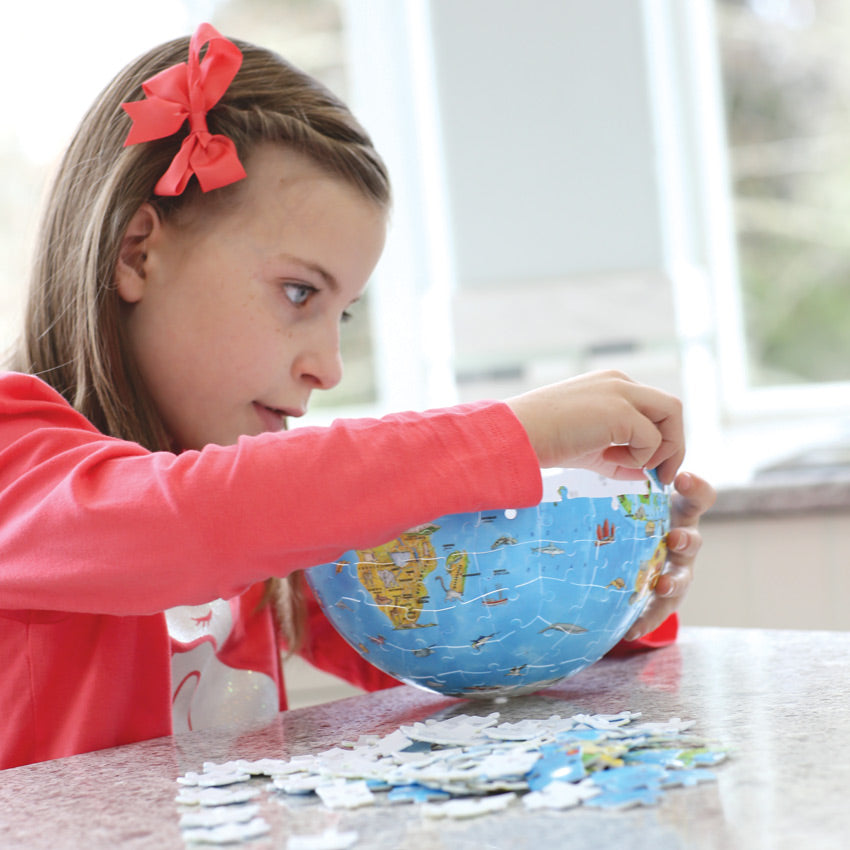 A brunette girl in a bright pink shirt and bow in her hair, is sitting at a kitchen counter and putting together the Puzzleball Globe. The 3 D globe is half finished while she is holding it up and putting a piece into place. The rest of the pieces are scattered in a pile on the countertop. The potion of the globe showing is of the bottom of Africa.
