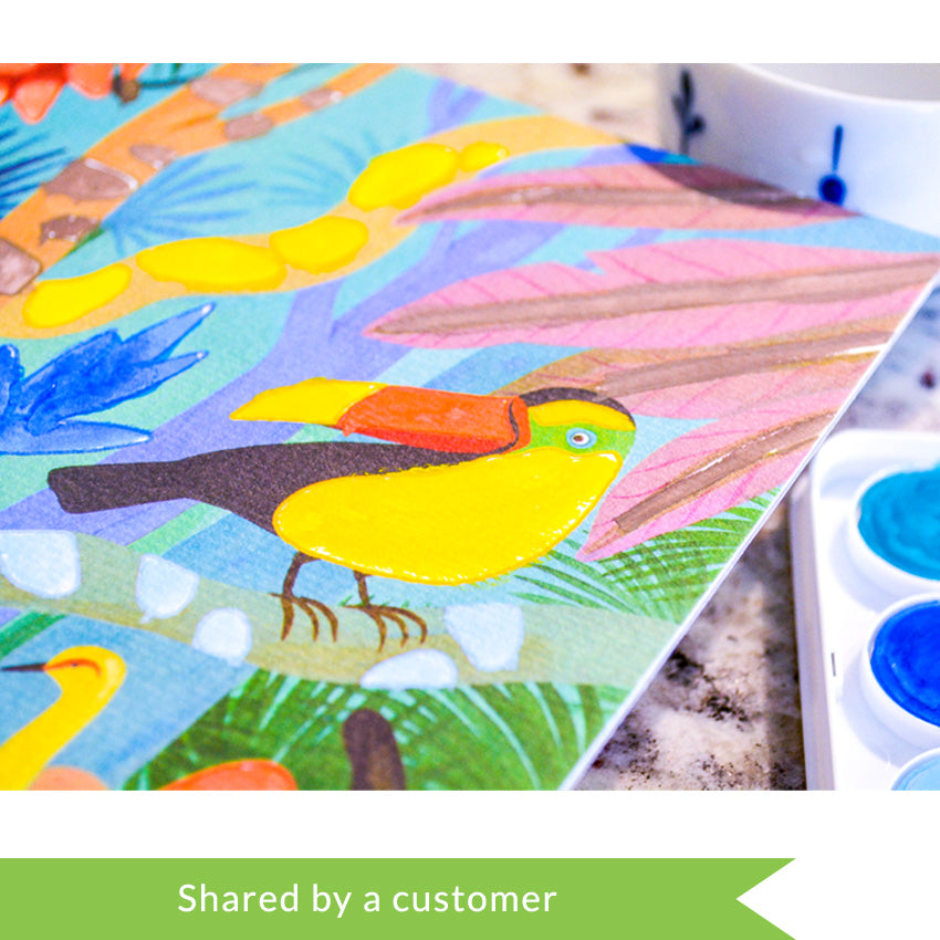 A customer photo of the Djeco Natural World Workshop jungle scene, laid out on a marble surface. The close-up shot focuses on a toucan surrounded by trees and bushes, in the colorful jungle scene. In the bottom-right you can see some blue paints in the tray and a tea mug with water in the upper-right corner.