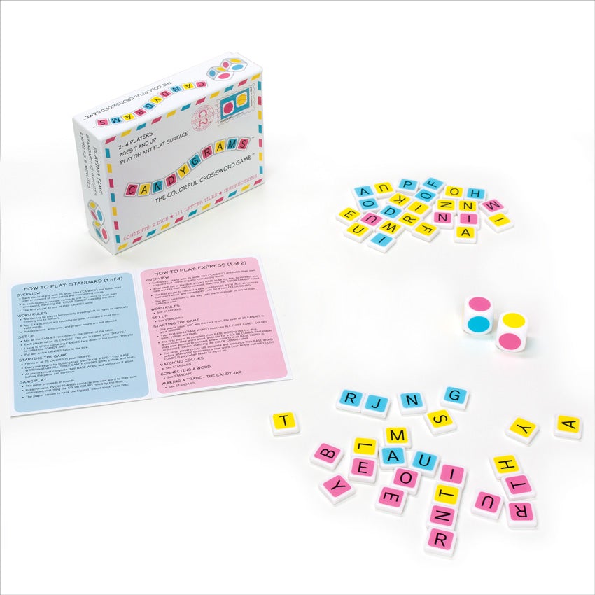 Candygrams game in play. The game box and instructions are on the left. To the right, on the top and bottom, are piles of colored letter tiles. The tiles are pink, yellow, and blue with white sides and backs. In between the piles on the right are 2 dice with alternating colored dots on each side, in red, yellow, and blue.