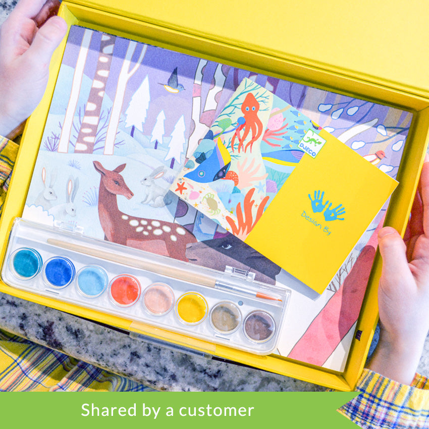 A customer photo of hands holding the yellow Djeco Natural World Workshop box open over a marble table. Inside the box, you see a winter scene with deer, rabbits, and snow-covered trees. Over the top is the instruction book and the paint tray with a paintbrush inside.
