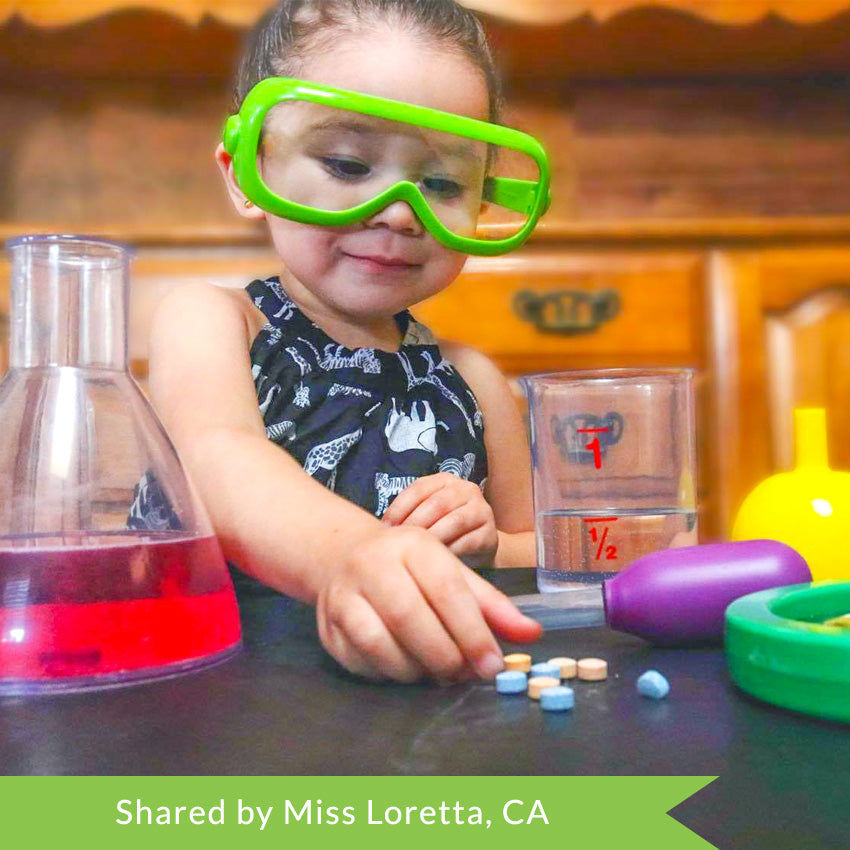 A customer photo of a small child with a slight grin in green safety goggles reaching out to grab a Color Fizzer tablet that is laying on the black table. On the table surrounding the child are child's lab equipment, including; a beaker, large eyedropper, measuring cup, funnel, and magnifying glass.