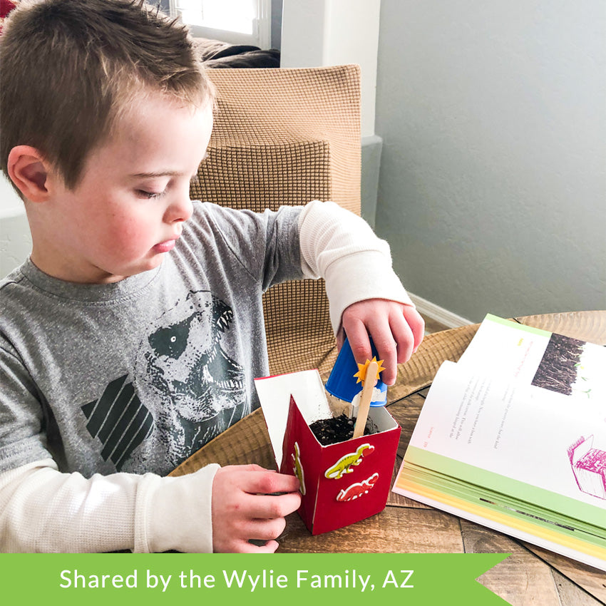 A customer photo of a young boy in a dinosaur shirt planting seeds into a maroon painted milk carton with dinosaur stickers. He has the What Your Preschooler Needs to Know book open for direction on the table in front of him.