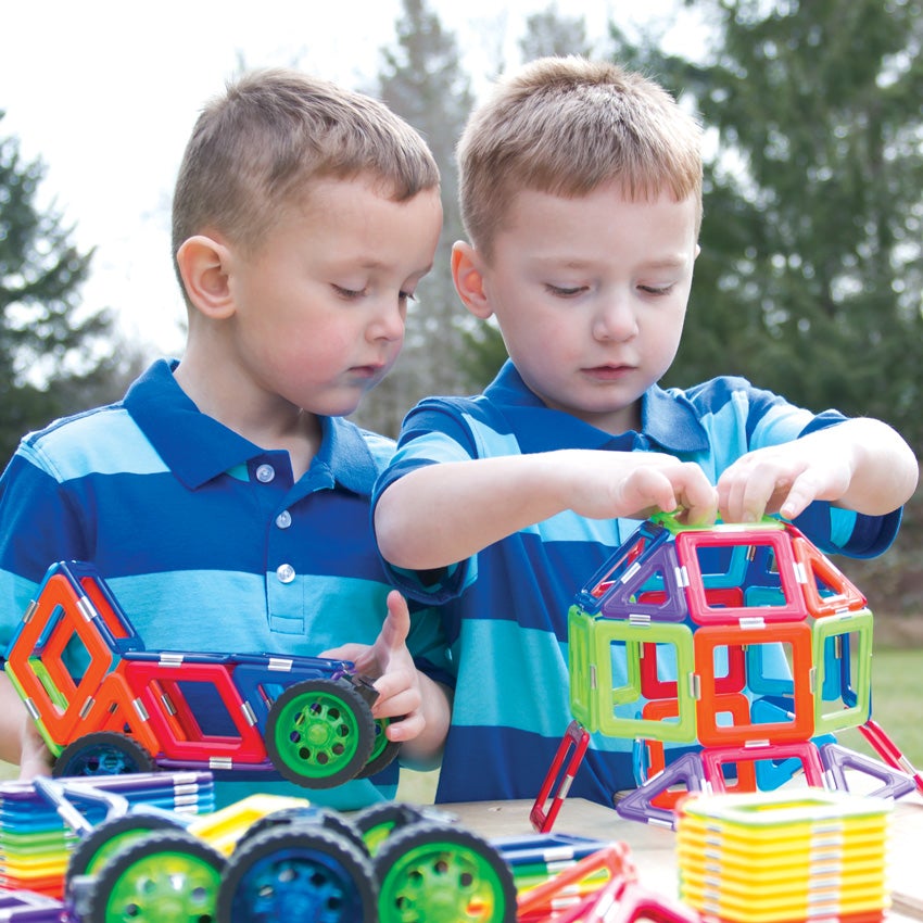2 blonde boys in striped shirts are outside and are putting together a large shape, made up of the GeoSmart Educational Deluxe pieces on a picnic table. The pieces are geometrical shapes in squares and triangles and are a variety of different colors.  The boy on the left is holding a car created by the magnetic pieces. The boy on the right is holding a large rounded object. There are pieces in stacks and piles on the table.