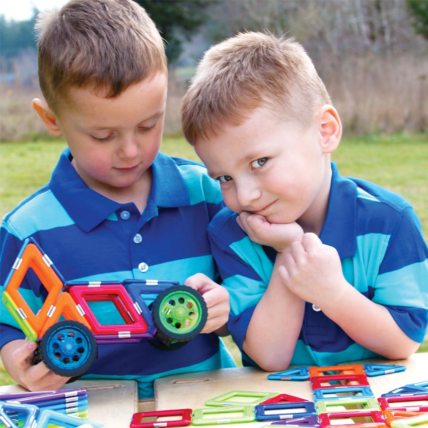 2 blonde boys in striped shirts are outside are playing with the GeoSmart Educational Deluxe set. The pieces are geometrical shapes in squares and triangles and are a variety of different colors.  The boy on the left is holding a car toy, made from the pieces. The boy on the right is leaning on his hands with his elbows on the table with a cheeky look on his face. There are pieces in piles and stacks on the table, along with a shape started by the boy on the right.