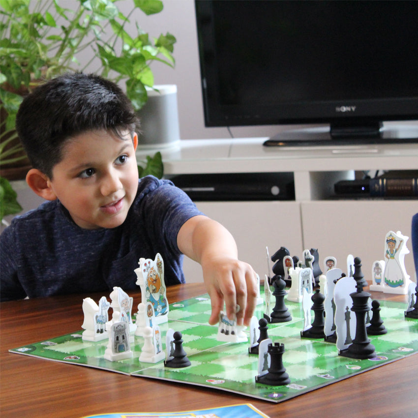 A dark-haired boy sits at a table with the Story Time Chess board in front of him. He has a slight smile and is reaching out his hand to move a piece on the board. He is looking up at someone across the table that you can’t see. The game board on the table has light and dark checkered squares with the game pieces in play.