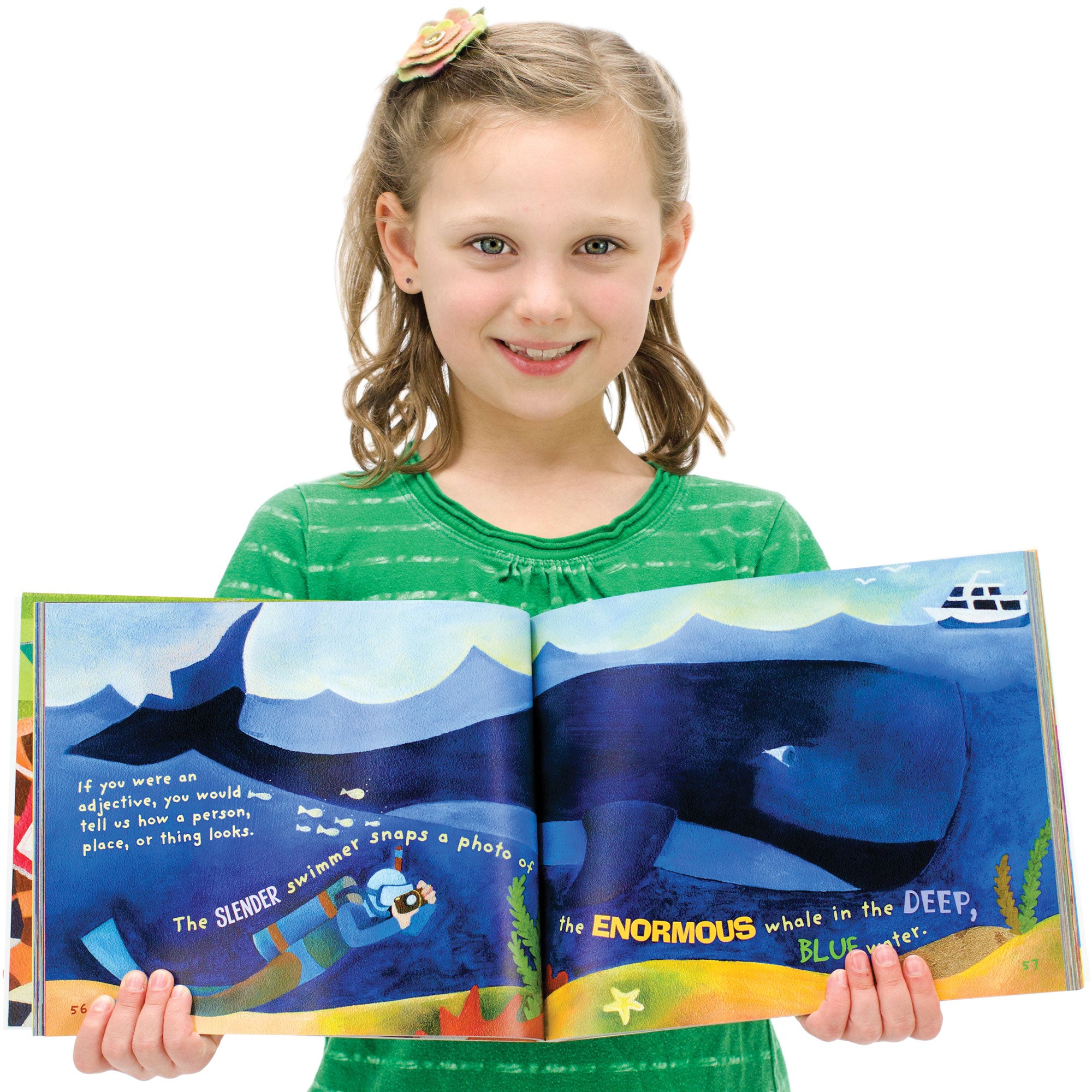 A young, blonde girl is smiling and holding open the Word Fun book to show inside pages. The pages show a large whale under the sea. There is a ship on top of the water, and a scuba diver under the water, taking a picture of the whale. The text on the pages reads “If you were an adjective, you would tell us how a person, place, or thing looks. The slender swimmer snaps a photo of the enormous whale in the deep, blue water.” The adjectives are large, bold, and colored differently than the rest of the text.