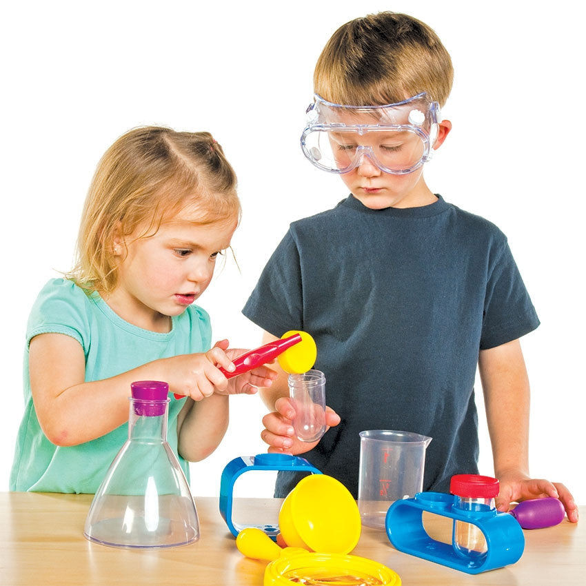 2 children playing with Primary Science Lab Kit items. The dark blonde girl on the left in a mint green shirt is holding large red tweezers grasping a yellow test tube cap held by a boy on the right. The dark blonde boy on the right is wearing safety goggles and a dark gray-blue shirt. In front of the children on the table are other contents of the Lab Kit.