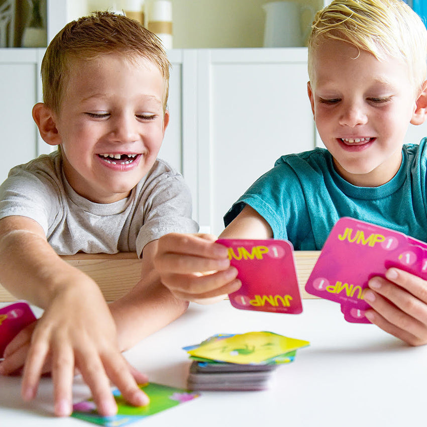 2 boys playing Jump 1. The boy on the left is smiling with missing a front tooth and reaching out for a card. The boy on the right is smiling and holding onto several cards.