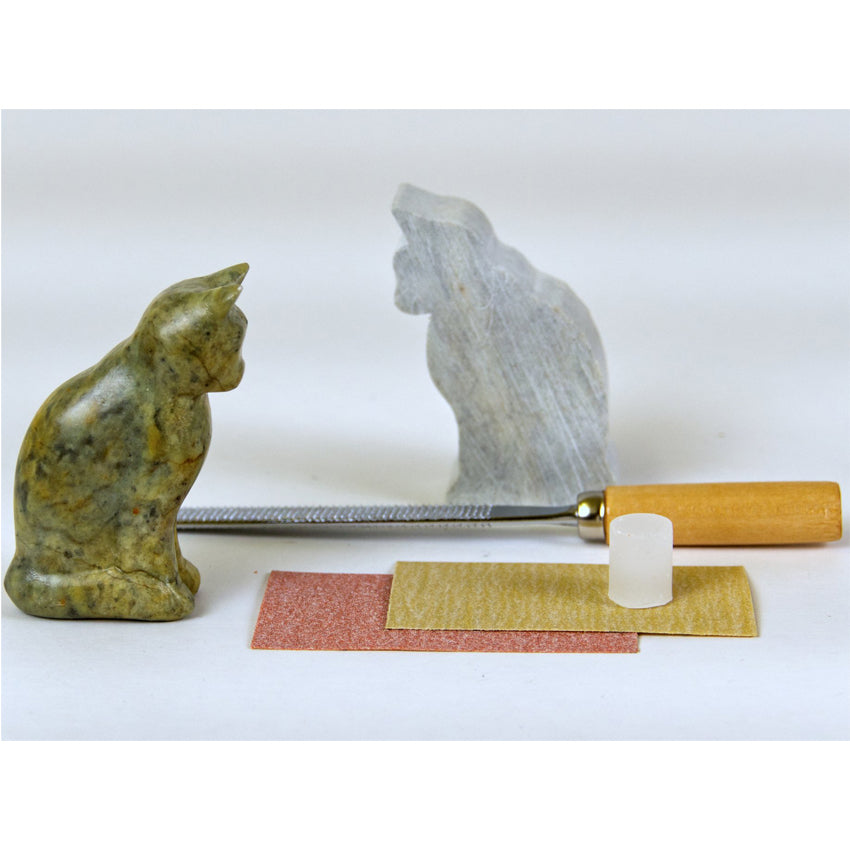 Cat Soapstone Carving Kit contents. On the left is the completed cat figure with a wood-handled metal file behind. Behind the file is a rough block-like cat figure. In front are 2 pieces of red and yellow sandpaper with a cylindrical chunk of wax on top.