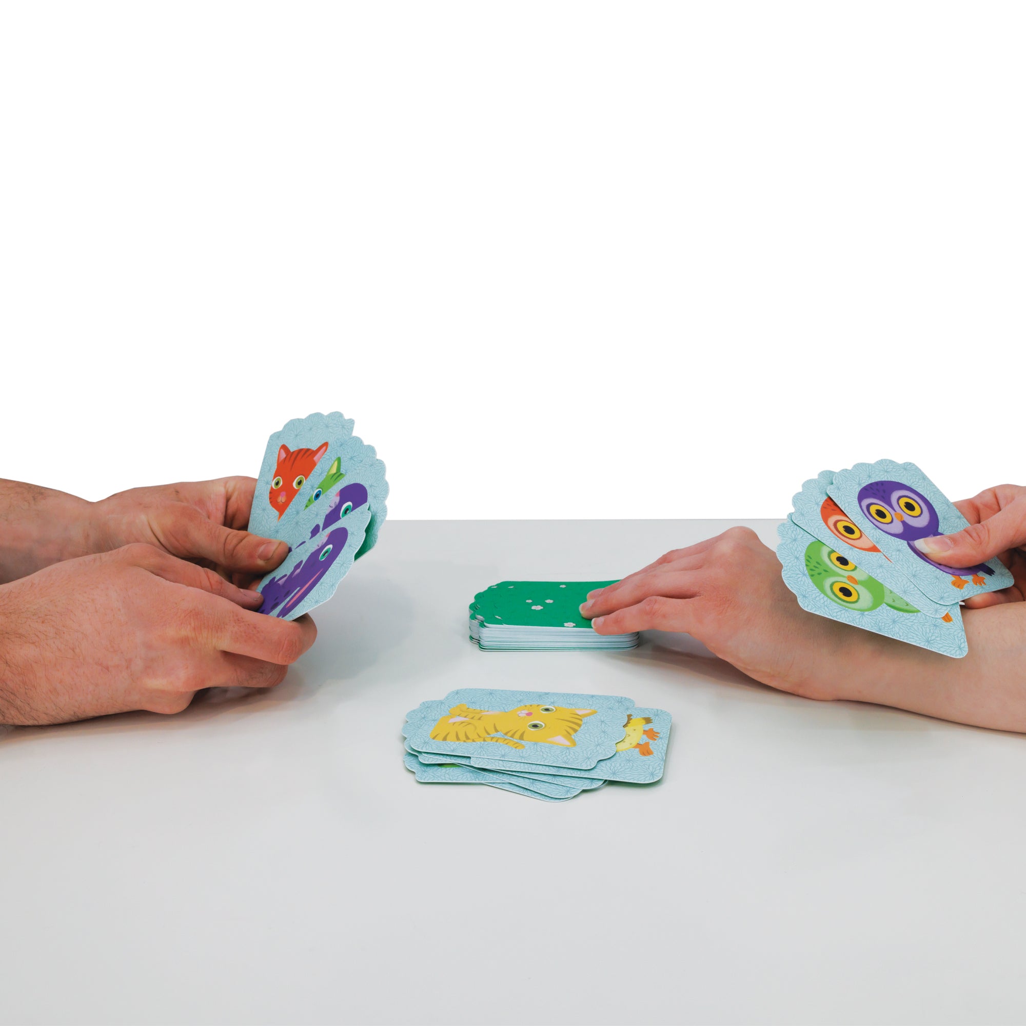 2 people’s hands on either side of the table playing the Djeco Little Match game. The left player is holding 4 cards. The right player is holding 3 cards, and is drawing another card with their left hand. There are 2 piles of cards in the middle; one stack face down, and one stack face up. The cards have different colored animals on each.