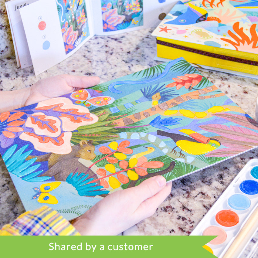 A customer photo of hands holding the Djeco Natural World Workshop jungle project over a marble table. In the top-left of the photo, you can see the instruction book open. In the top-right is the yellow product box with an underwater scene on the top. In the bottom-right, you can see the paint tray with a paintbrush inside.