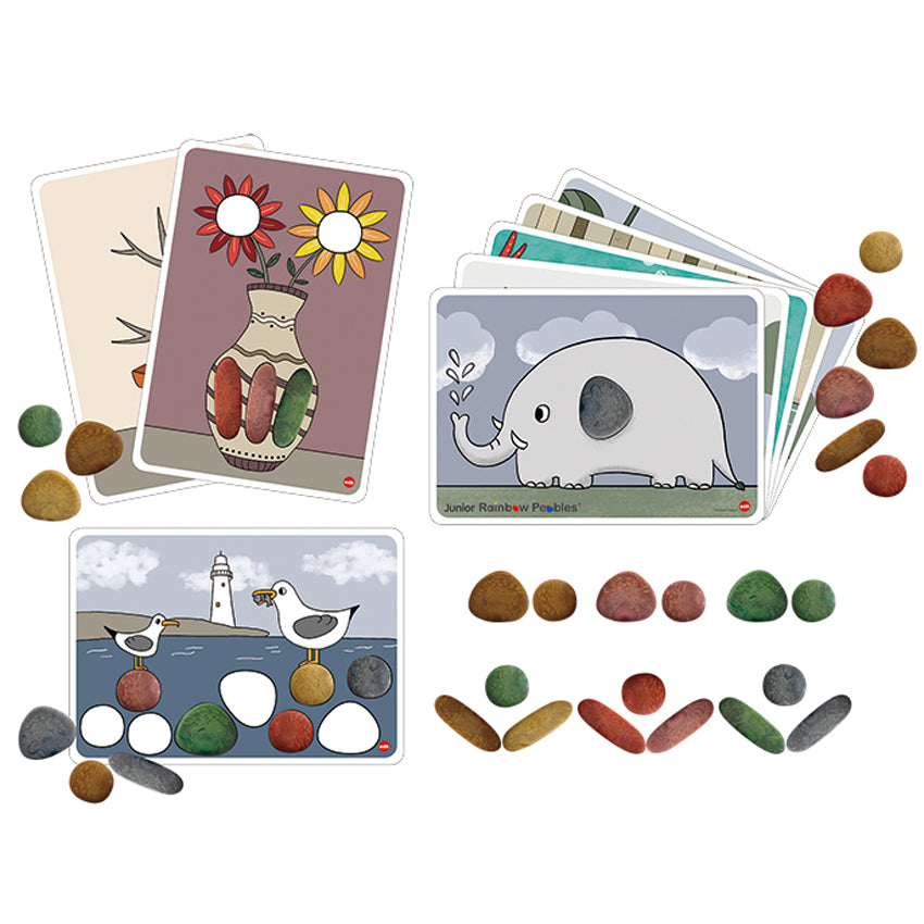 Junior Rainbow Pebbles, Earth Tones, contents spread out over a white surface. The activity cards have illustrations with blank spaces to put the pebbles. The cards on the left show a vase with flowers and a lighthouse beach scene with seagulls. The card the right shows an elephant spraying water in the air. There are more cards stacked below the visible ones. There are pebbles, in many shapes and colors, spread over the surface and cards.