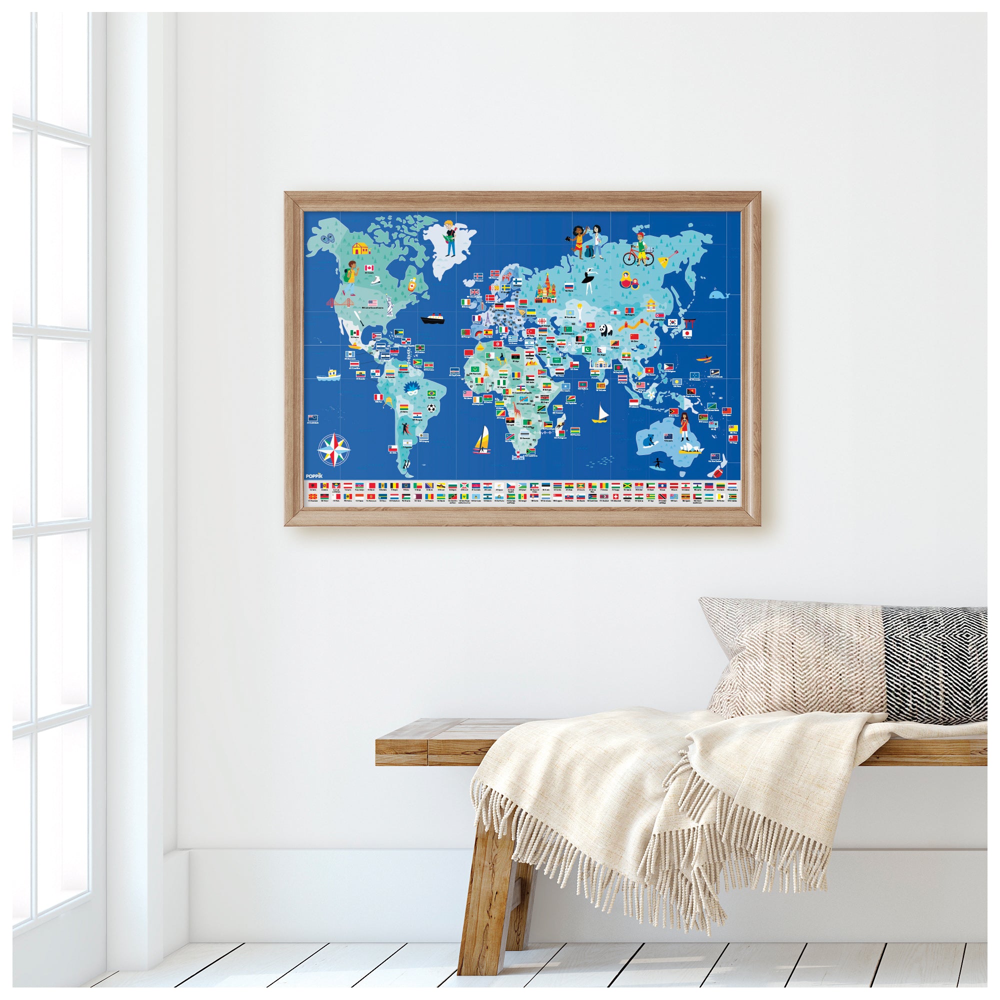 World map with flags framed and hung on a white wall. Under the map is a bench with an ivory colored blanket and a pillow placed on top of a white floor.