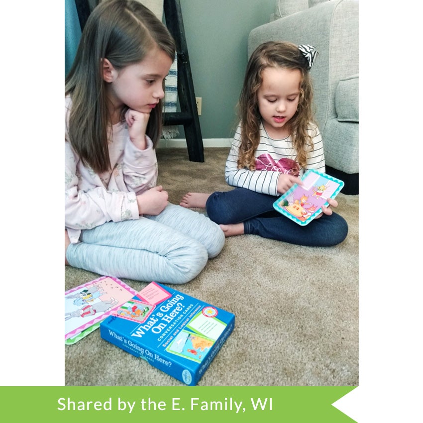 A customer photo of 2 brunette girls playing with the What’s Going on Here Conversation Cards. The older girl on the left is sitting with her legs folded under her and is paying attention to the girl on the right who is explaining the card she is holding. The card box and 2 cards are sitting on the floor in the bottom-left of the picture.
