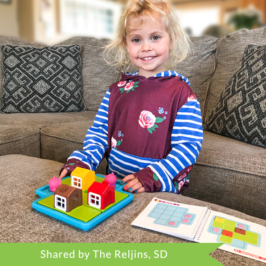 A customer photo of a young, smiling, blonde girl playing with the Three Little Piggies game. She is kneeling with a couch behind her and playing the game on the ottoman in front of her. The game board is light blue with playing pieces on the board, including; a red house, a yellow house, 3 pigs, and a wolf. The instruction booklet, off to the right, is open to a challenge page.