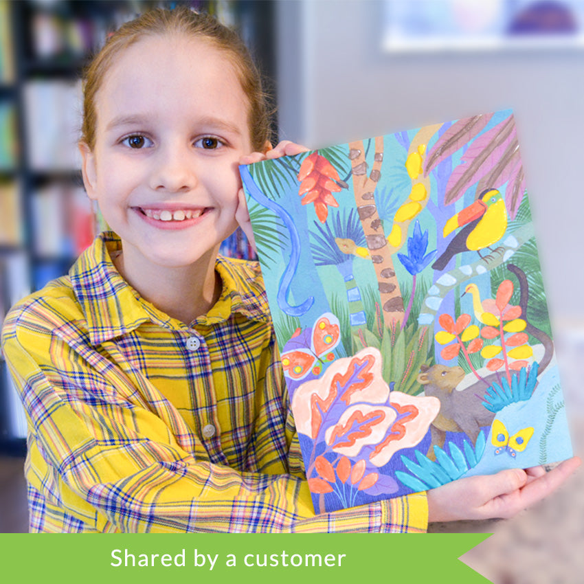 A customer photo of a dark strawberry-blonde girl holding up the Djeco Natural World Workshop jungle project and smiling big. The jungle scene has many trees and bushed in a variety of colors .There is also a toucan, snakes, butterflies, and a few other jungle creatures. 