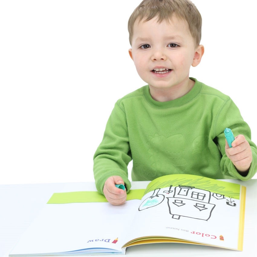 A boy in a green sweatshirt is sitting at a white table holding a green coloring tool in his left hand and the cap to the coloring tool in the right hand. On top of the table is the Draw + Learn Animals + Places book, opened to show a house doodle, grass, and a tree that has been partially colored in green.