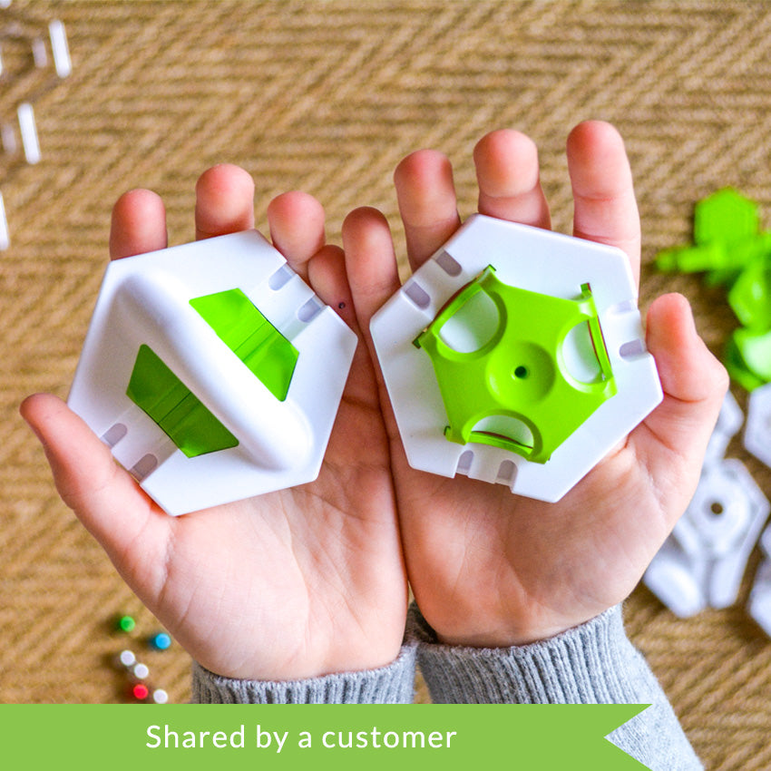 A customer photo from the top of a child’s hands holding 2 white hexagon pieces with bright green obstacles in the middle from the GraviTrax set. Below the hands and out of focus are playing pieces from the set. The pieces shown, but out of focus, are white hexagons, green hexagon attachment pieces, colored marbles, and a clear hexagon plate. 