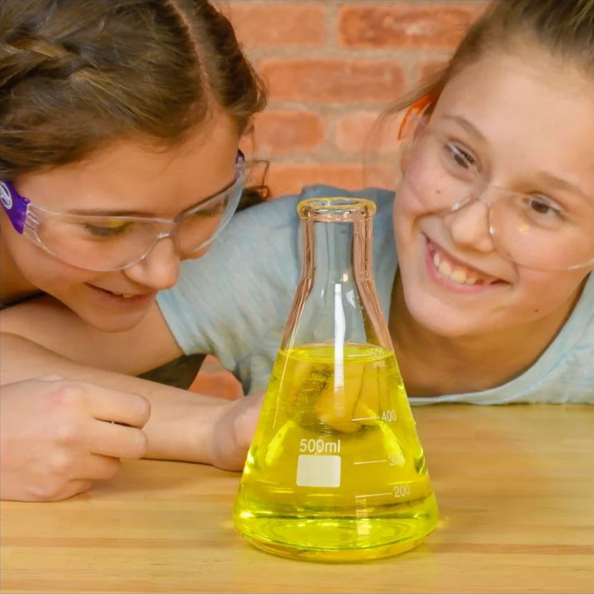 Two girls with safety goggles watch with smiles as Color Fizzers color a beaker with water in front of them. The beaker, that is sitting on a wood table, has yellow water with a yellow tablet coloring the water.