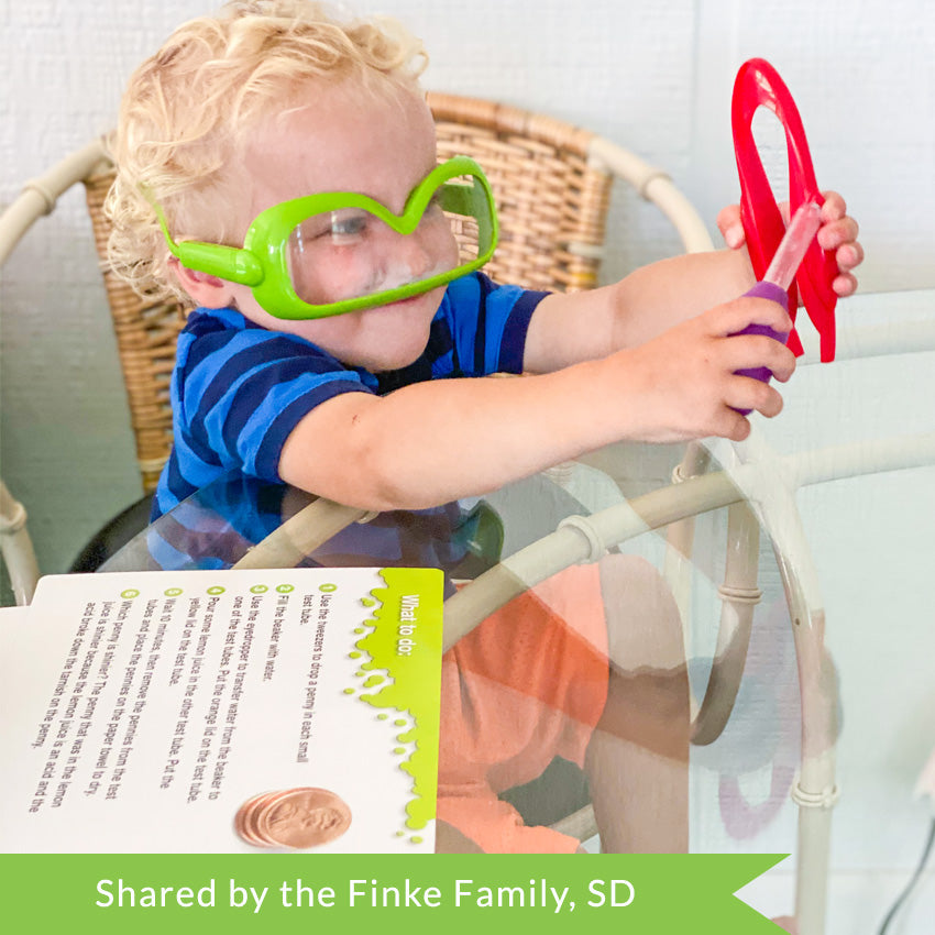Customer photo of a blonde toddler wearing green safety goggles upside down and a blue striped shirt.  He is holding large red tweezers in his left hand and a eyedropper in his right hand. On the glass table in front of him are the activity cards from the Primary Science Lab Kit.