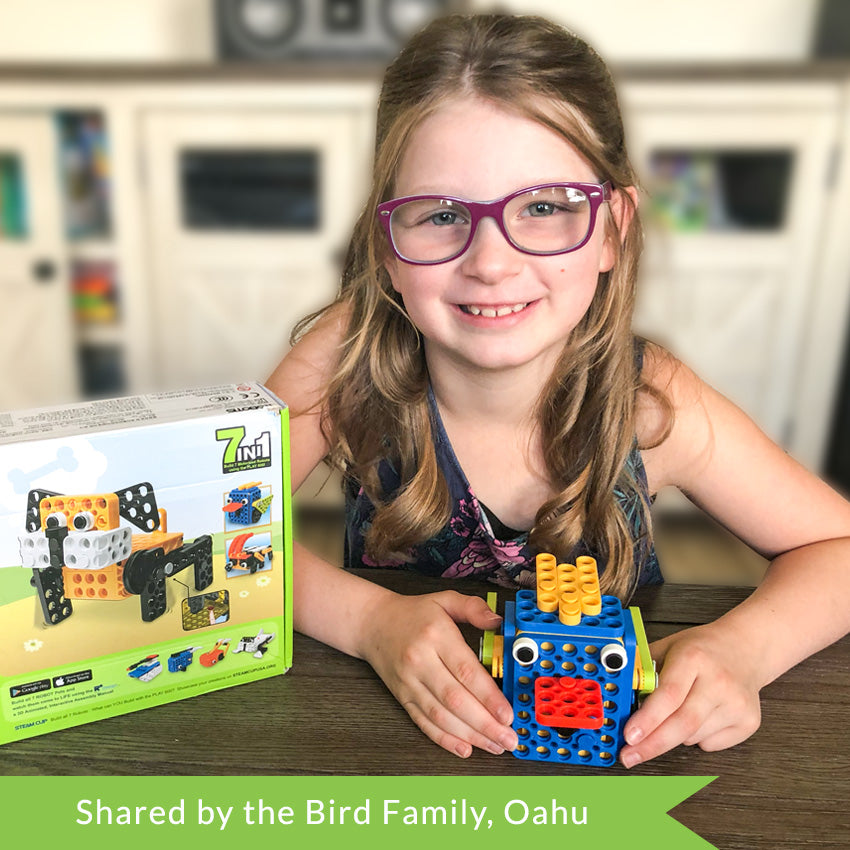 A customer photo of a young blonde girl with purple glasses holding a bluebird robot on top of a dark wood table. To the right of her is the box for the Robotis Play 600 Pets with a dog robot on the cover.