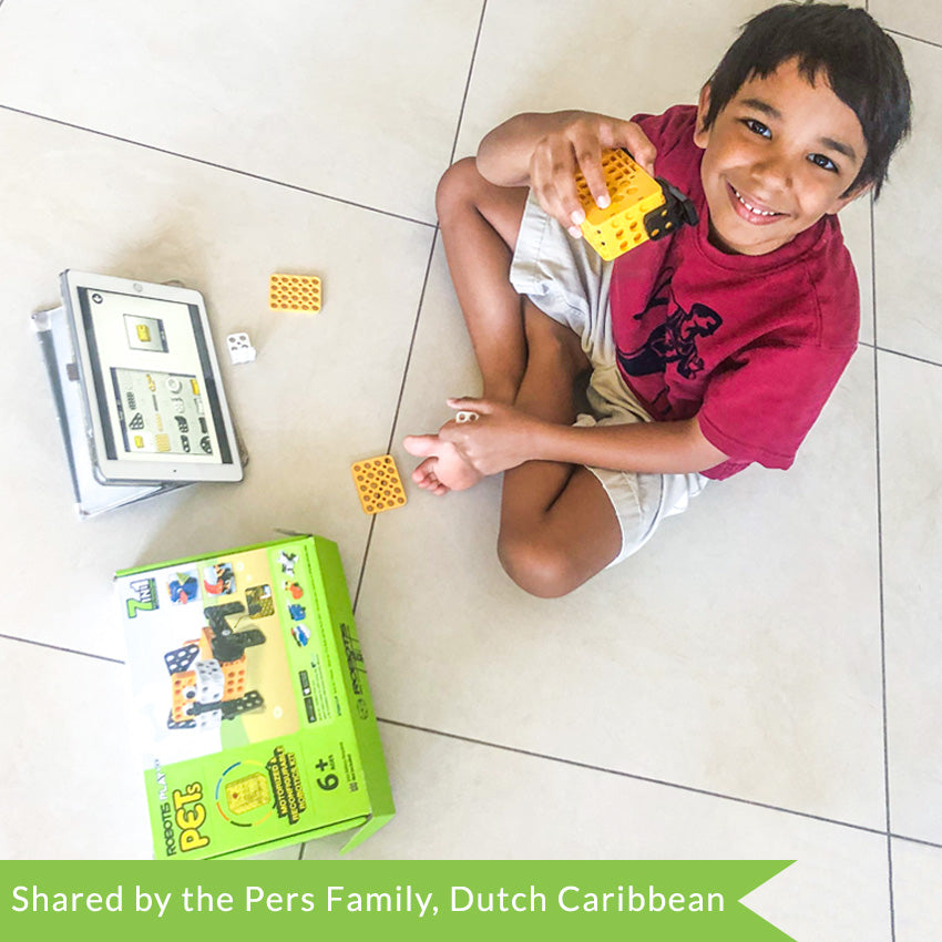 A customer photo of a smiling black-haired boy sitting cross-legged and holding the motor piece of the Robotis Play 600 Pets up toward the camera.  On the tile floor he is sitting on is a tablet with build instructions and the product box with a few scattered pieces.