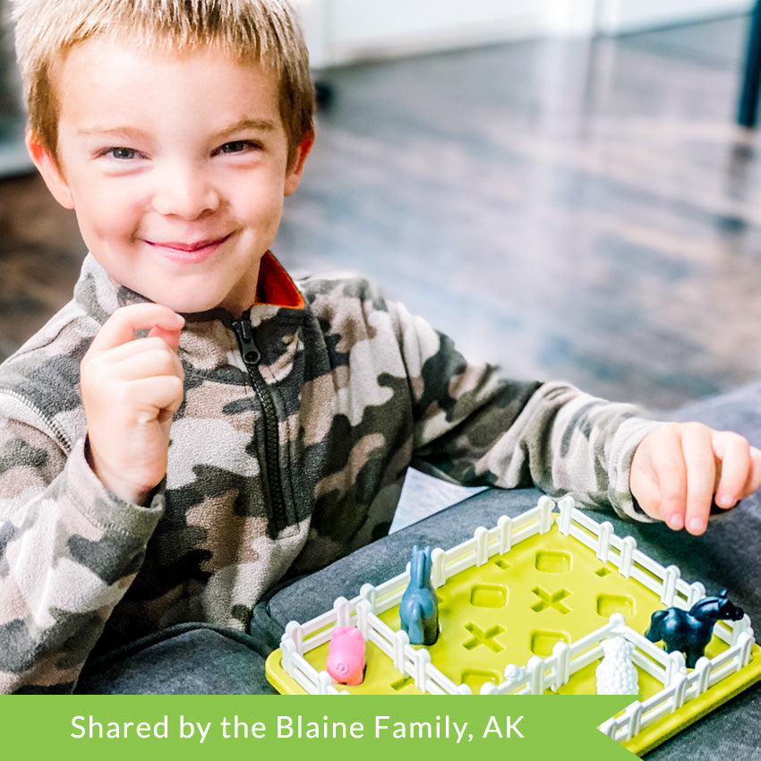 A customer photo of a young blonde boy in a camouflage jacket, smiling, and playing with the Smart Farmer game. He has his hands in the air waiting to arrange pieces on the board. The green rectangle-shaped game board has white fence pieces all around the edge and a few through the middle. You can see a pig, sheep, and 2 horses inside the fenced area.